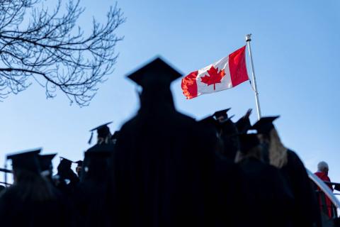蓝天上飘扬着一面加拿大国旗，前景中是UBC 2022届毕业生戴着帽子、身穿长袍的身影。开云体育要一倍流水